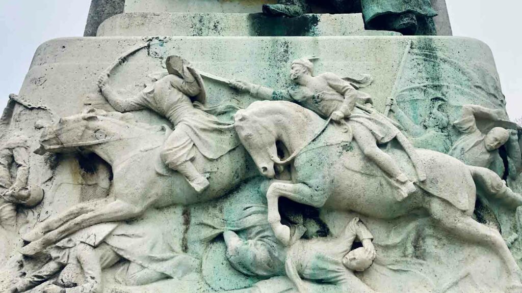 Fotografía de una escultura de piedra en Budapest, Hungría, que representa una batalla en la que un hombre decapita la cabeza de otro jinete a caballo en combate.